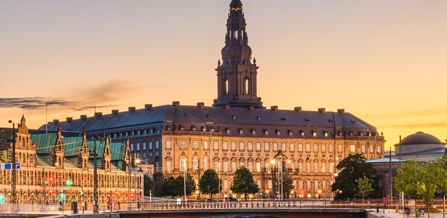 A historic European building in Copenhagen, graced by a central tower at sunset, stands surrounded by trees and streetlights, casting an enchanting glow that feels like stepping into a timeless session.