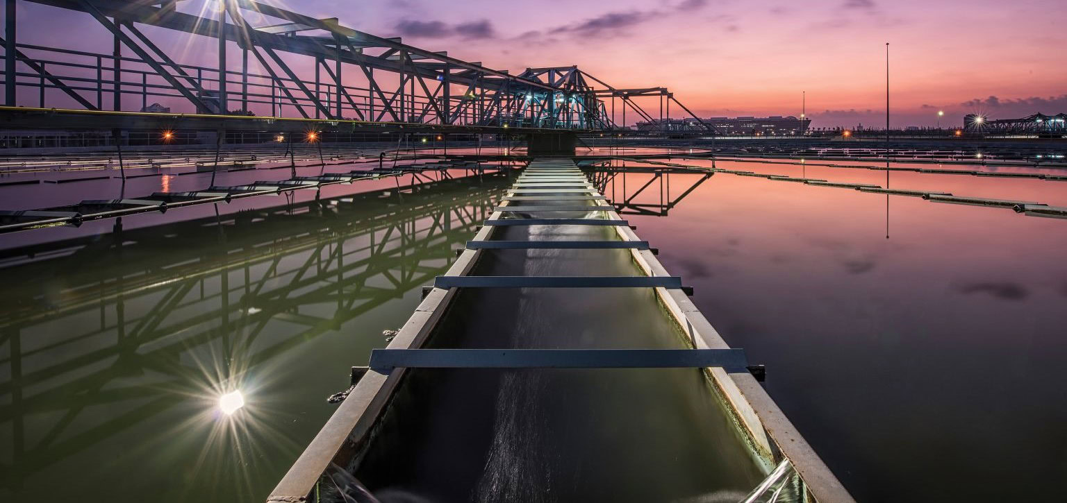 A long bridge-like structure spans across a calm water body at dusk, with a reflection of the bridge and purple sky on the water's surface, resembling a serene setting for a Digital Leadership Summit.