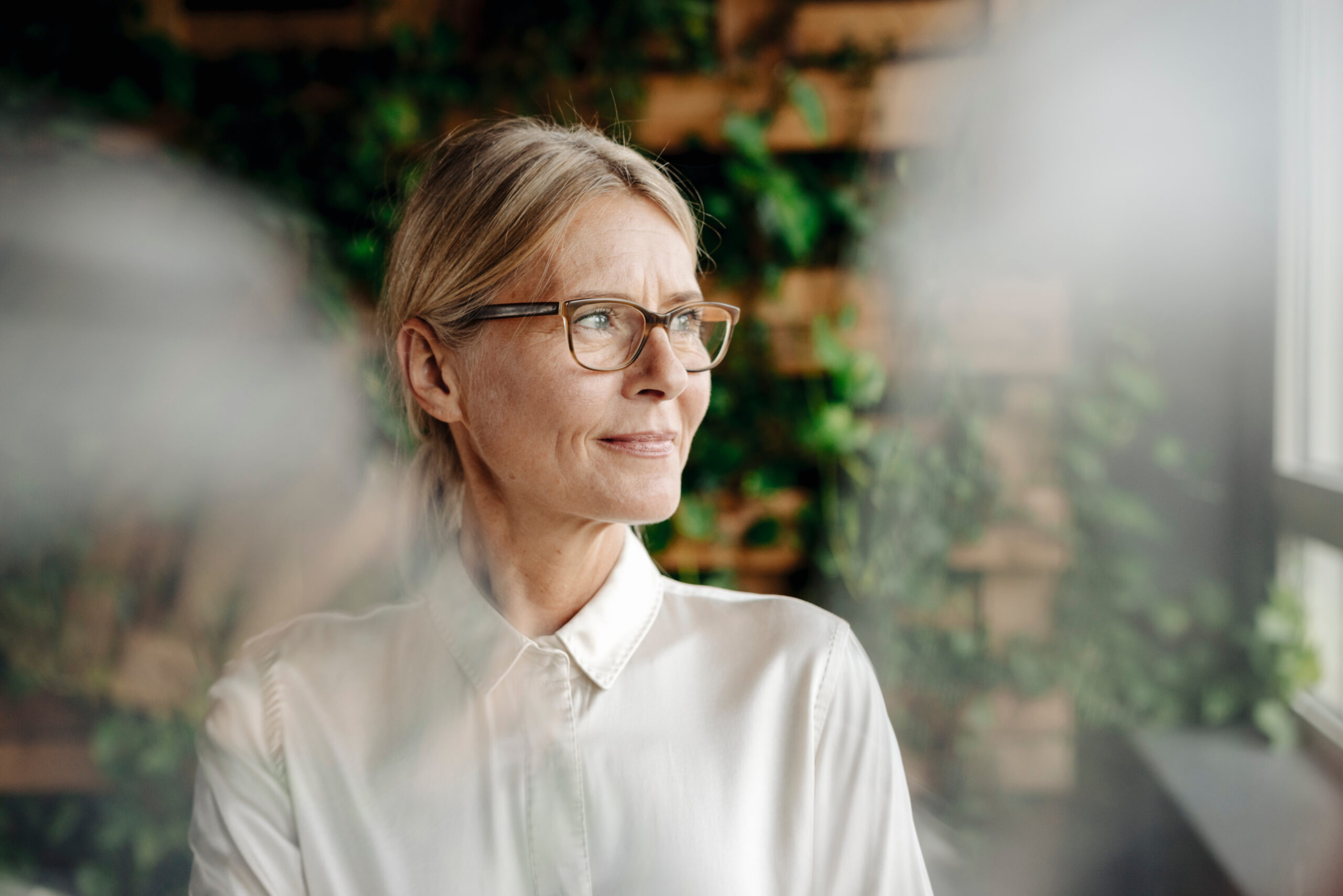 An older woman with glasses and a white shirt gazes out a window, with a background of green foliage and brick.