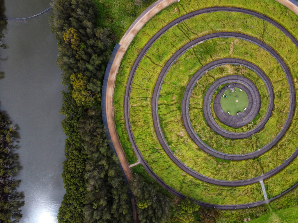 Aerial view of a circular, terraced garden with grassy concentric rings. A river runs alongside the greenery, bordered by dense trees.