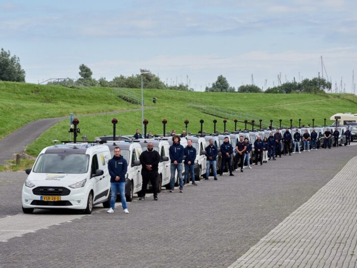 People standing in front of a line of identical white vans equipped with AI-Infra Solutions roof-mounted cameras, parked on a paved area near a grassy embankment in the Netherlands.