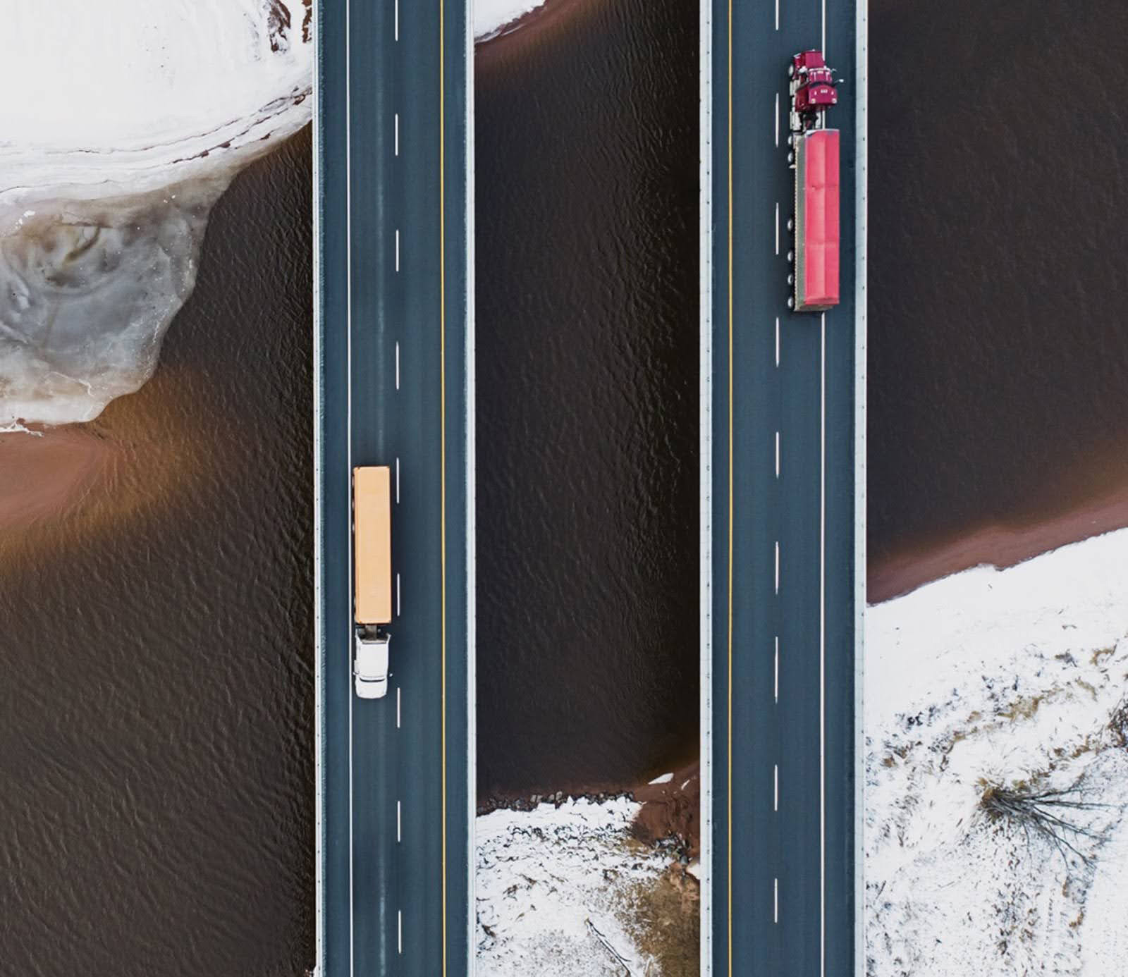 Aerial view of two trucks, winners on their journey, traveling on parallel highways over a river, with a snowy landscape surrounding them.