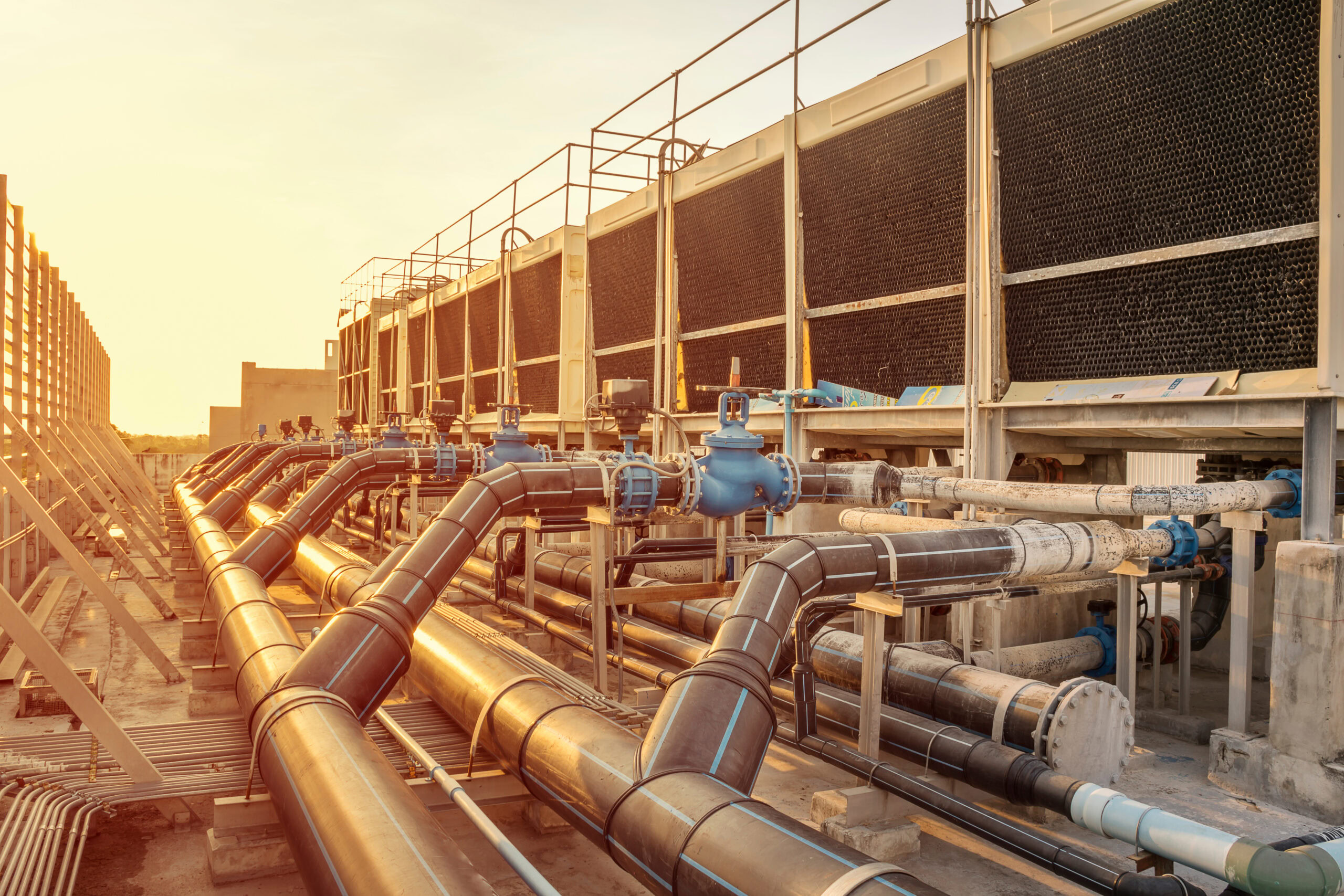 Industrial facility with large metal pipes and blue valves arranged in a complex network, resembling an Auto Draft system, situated next to large gray cooling towers at sunset.