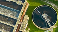 Aerial view of a wastewater treatment plant featuring a rectangular aeration basin on the left and a circular clarifier on the right, expertly managed through professional services, all surrounded by lush green grass.