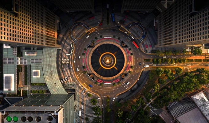 Aerial view of a circular urban intersection with surrounding tall buildings and lush greenery. Traffic flows smoothly around the central structure, with roads and crosswalks radiating outward.