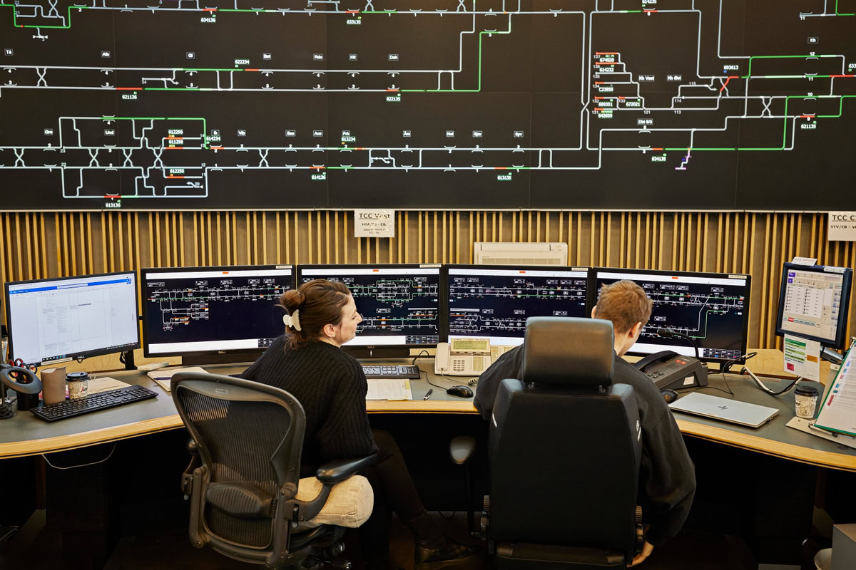 Two people are seated at workstations, monitoring multiple computer screens displaying complex data and diagrams of a railway system, with a large schematic map on the wall behind them.