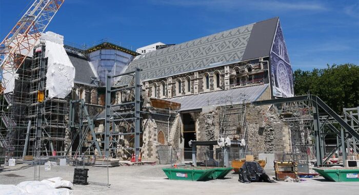 Construction and restoration work on the large, historic Christ Church Cathedral features scaffolding and cranes at the site. In the foreground, green dumpsters and building materials surround elegant stone columns, highlighting the grandeur of this architectural gem.