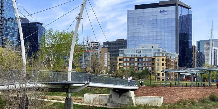 Nestled in a city park, the pedestrian bridge offers a serene stroll with Denver's skyscrapers, including a towering Hilton, as the backdrop and trees gracefully lining the path.