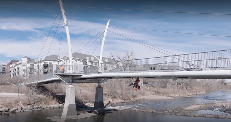 In Denver, a person in climbing gear hangs from a bridge over a river, while another in orange stands confidently on the bridge. Buildings and trees paint the backdrop of this adventurous user story.