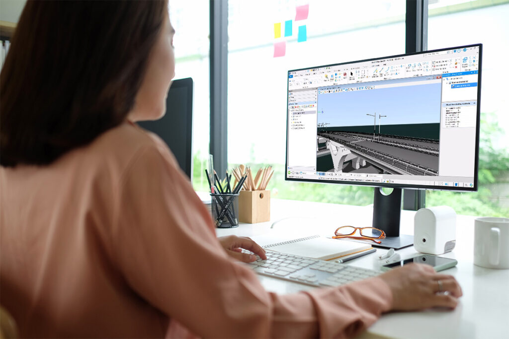 A user works on 3D modeling software, likely MicroStation, on a desktop computer. The screen displays a meticulously crafted 3D bridge design for 2024.