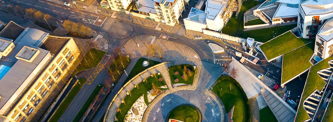 Vue aérienne d'une zone urbaine arborant une place circulaire avec un jardin, des bâtiments aux toits couverts de verdure, des rues et des voitures garées.