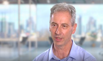 Man in a blue shirt discusses CaddPro solutions in an office setting, with the blurred cityscape of Australia as a backdrop, capturing the essence of our Partner Spotlight series.