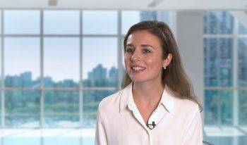 A woman in a white blouse with a clip-on microphone sits in a modern office space with large windows overlooking the cityscape, preparing for the Partner Spotlight segment.