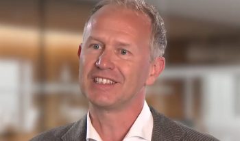 A middle-aged man with short, light hair and a light complexion is smiling, wearing a white shirt and grey blazer. Featured in our Partner Spotlight, he stands before a blurred indoor background representing AI-Infra Solutions.