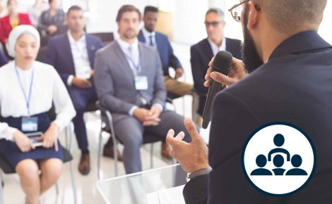 A speaker addresses an audience seated in a conference room, emphasizing important keywords throughout his talk. Holding a microphone and gesturing with his hand, he ensures the message is clear. Audience members in business attire, some with lanyard name tags, listen attentively to learn valuable insights.