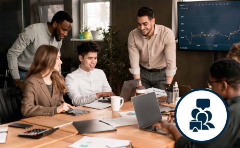 A diverse group of colleagues gathered around a conference table, learning together as they work on laptops and discuss a presentation displayed on a screen.