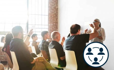 An instructor delivers a presentation to a seated audience in a bright room with large windows and brick walls. An icon featuring three people's heads in a circle with a curved line is in the bottom right, highlighting the learn-focused session that addresses SEO principles.