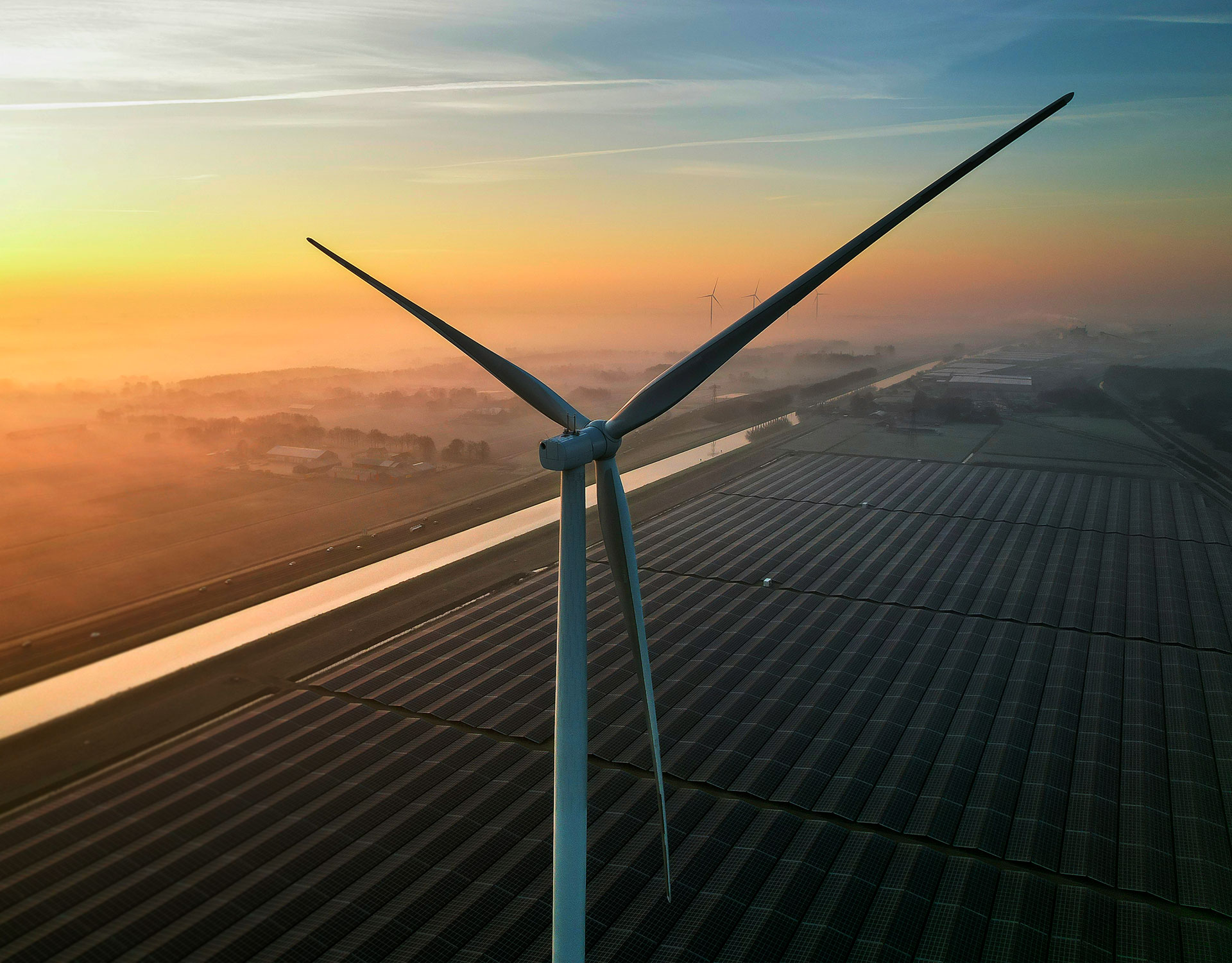 A wind turbine towering over solar panel fields during sunrise, with a canal and misty farmland in the background.