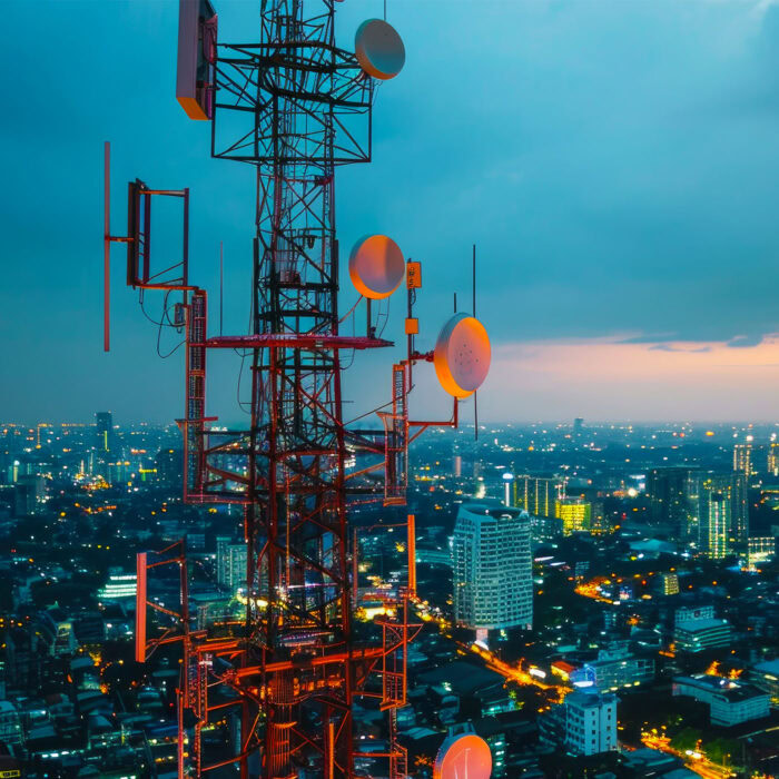 A telecommunications tower with multiple antennas stands against a cityscape at dusk, with lights from buildings and streets illuminating the background, a testament to the intricate network managed by Genesys International.