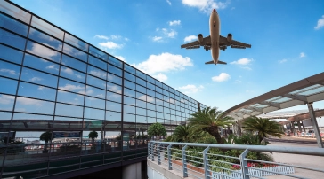 유리 외관과 푸른 하늘, 야자수가 있는 현대적인 공항 터미널 상공을 비행하는 비행기.