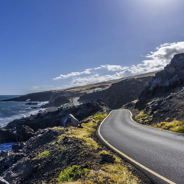 Eine kurvenreiche Straße entlang einer felsigen Küstenlandschaft mit dem Meer auf der linken Seite und einem strahlend blauen Himmel darüber, der teilweise von weißen Wolken bedeckt ist. Diese Ansicht könnte ein Beispiel für die Verwaltung durch das Verkehrsministerium von Hawaii sein.