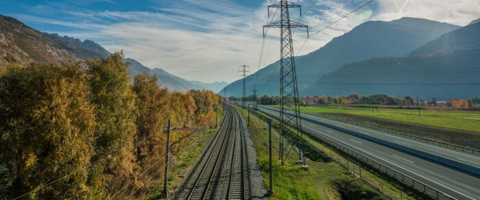 Railway tracks run parallel to a highway through a scenic mountain valley, flanked by autumnal trees and framed by towering power lines under a clear sky.