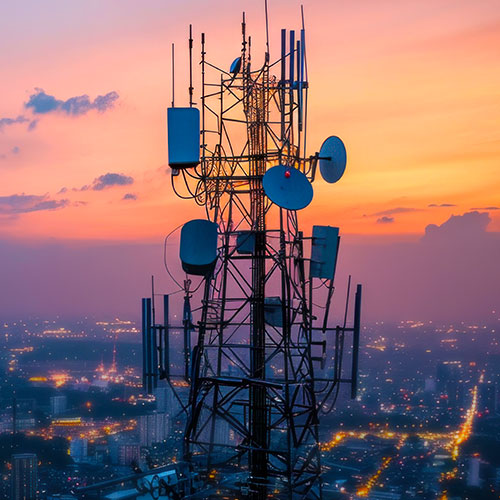 Ein Sendemast mit verschiedenen Antennen erhebt sich vor einer Stadtlandschaft im Dämmerlicht, im Hintergrund sind beleuchtete Straßenlaternen und Gebäude zu sehen.