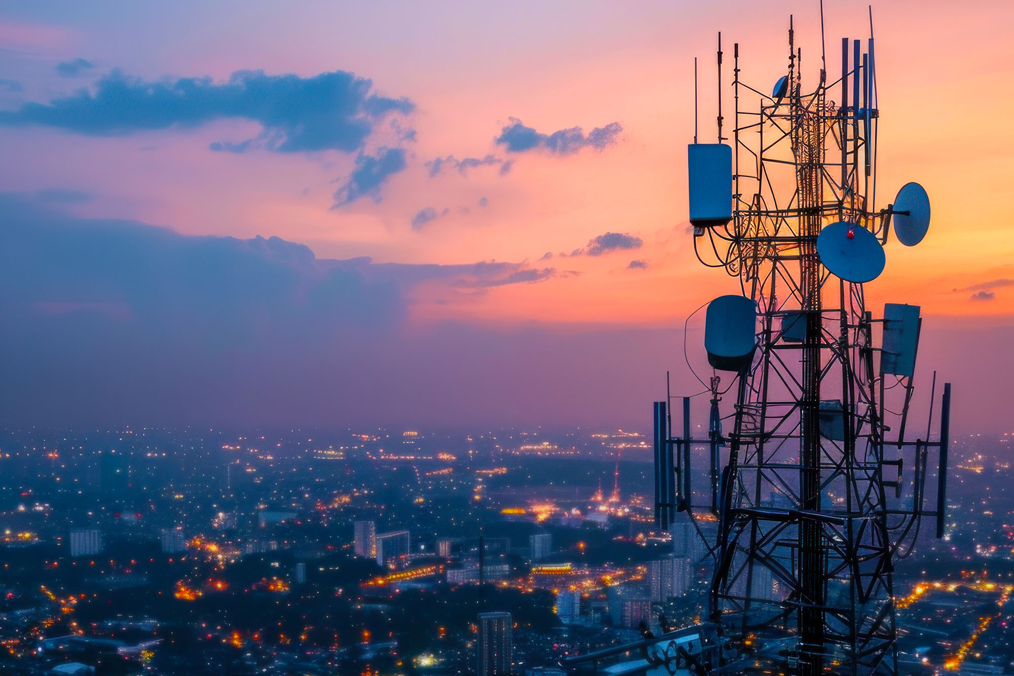 A communications tower with a sleek, modern design and multiple antennas stands against a vibrant sunset, overlooking a cityscape with illuminated buildings.