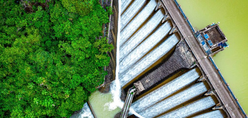 Aerial view of a dam with water flowing over spillways adjacent to a dense forested area, capturing the essence of Infrastructure 2024.