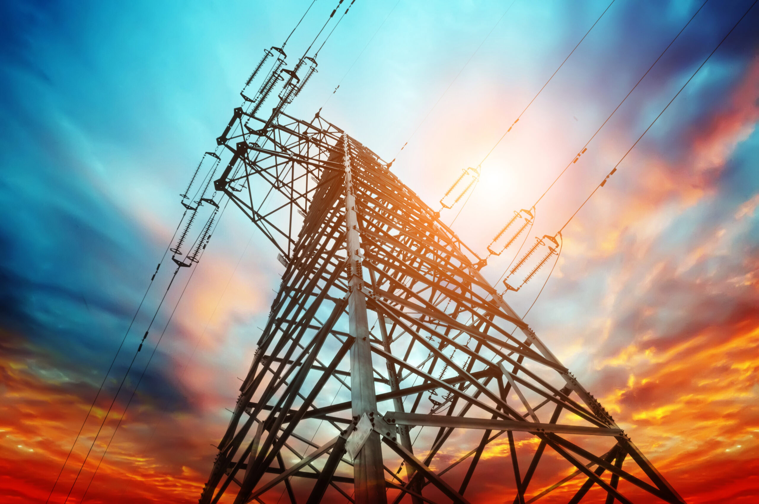 A tall metal electricity transmission tower extends into a colorful sky at sunrise or sunset, embodying the spirit of our Energy Edition, with power lines stretching outward.