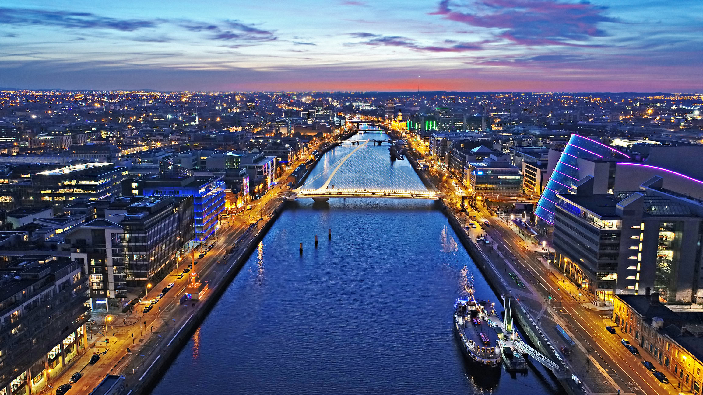 Aerial view of Dublin at dusk, featuring a river with illuminated bridges, modern buildings on both sides, and a colorful sky in the background. The cityscape comes alive as if in perfect harmony, reminiscent of SYNCHRO Sessions.