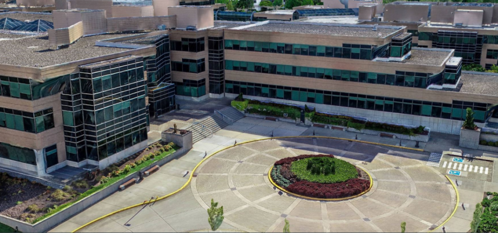 Vista aérea de um moderno complexo de escritórios com um jardim circular no pátio da frente, cercado por caminhos pavimentados e vagas de estacionamento, capturada usando serviços avançados de nuvem.
