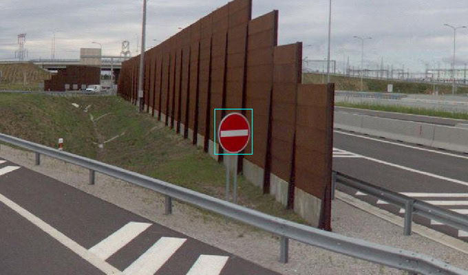 A "No Entry" sign is posted beside a highway with sound barriers and a cloudy sky in the background.