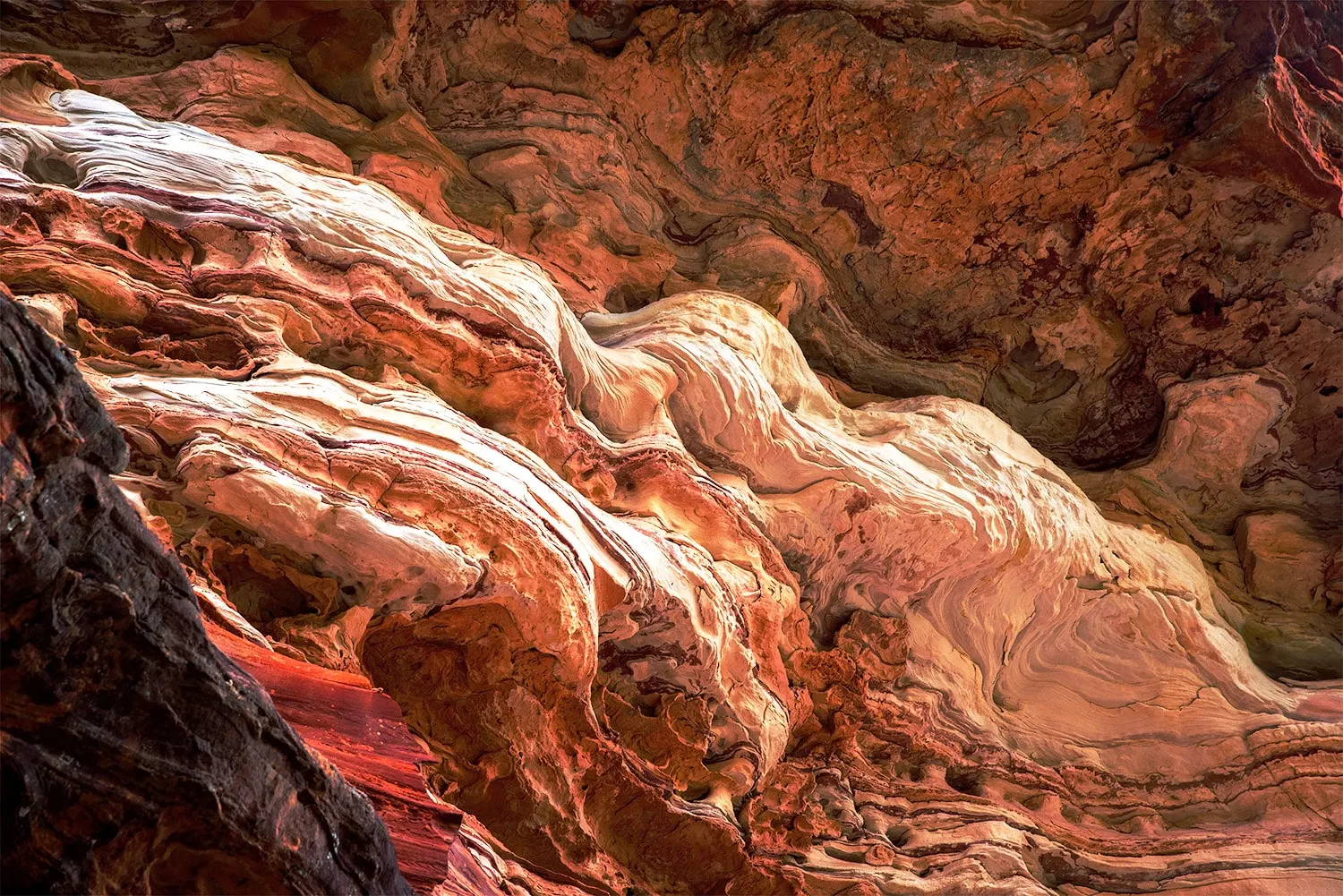 A detailed view of swirling, layered rock formations with shades of red, orange, and beige, resembling waves or folds.