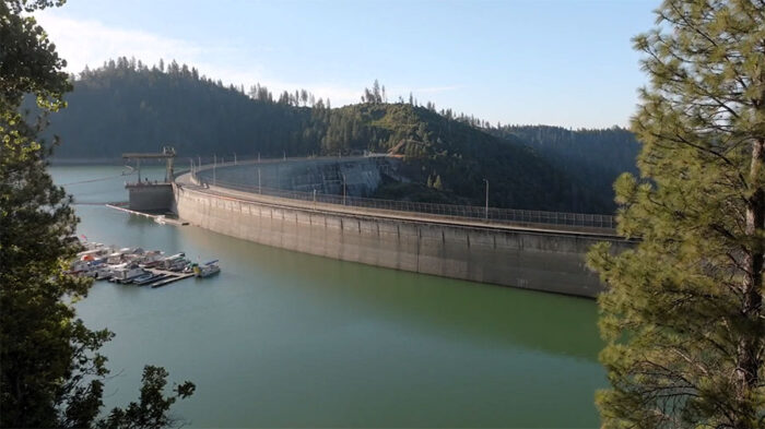 A large concrete structure, like Bullards Bar Dam, stretches across a serene body of water, surrounded by forested hills under a clear sky.