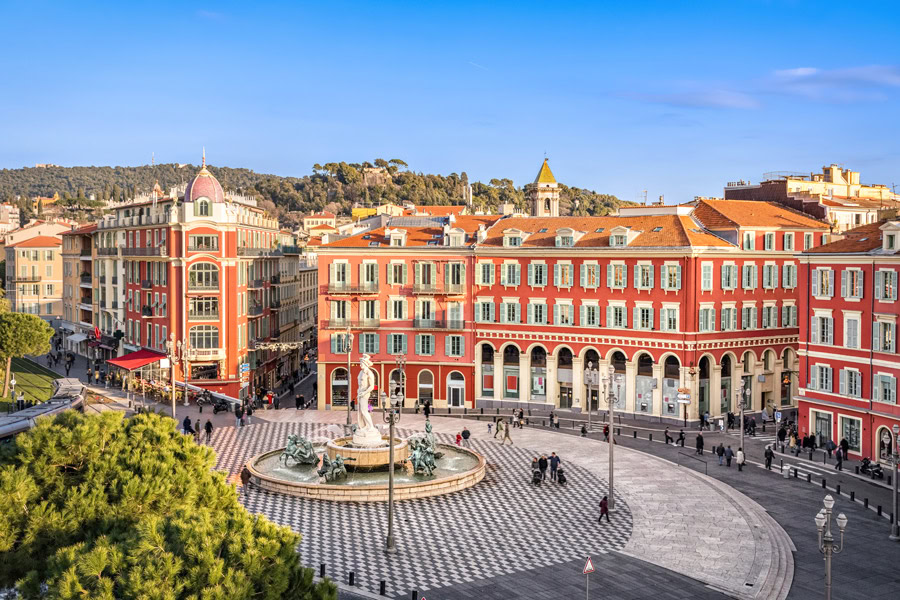 A town square with a central fountain surrounded by colorful buildings and a charming hotel. People are walking and gathering around the square. Trees and a hill can be seen in the background under a clear blue sky in Nice, France.
