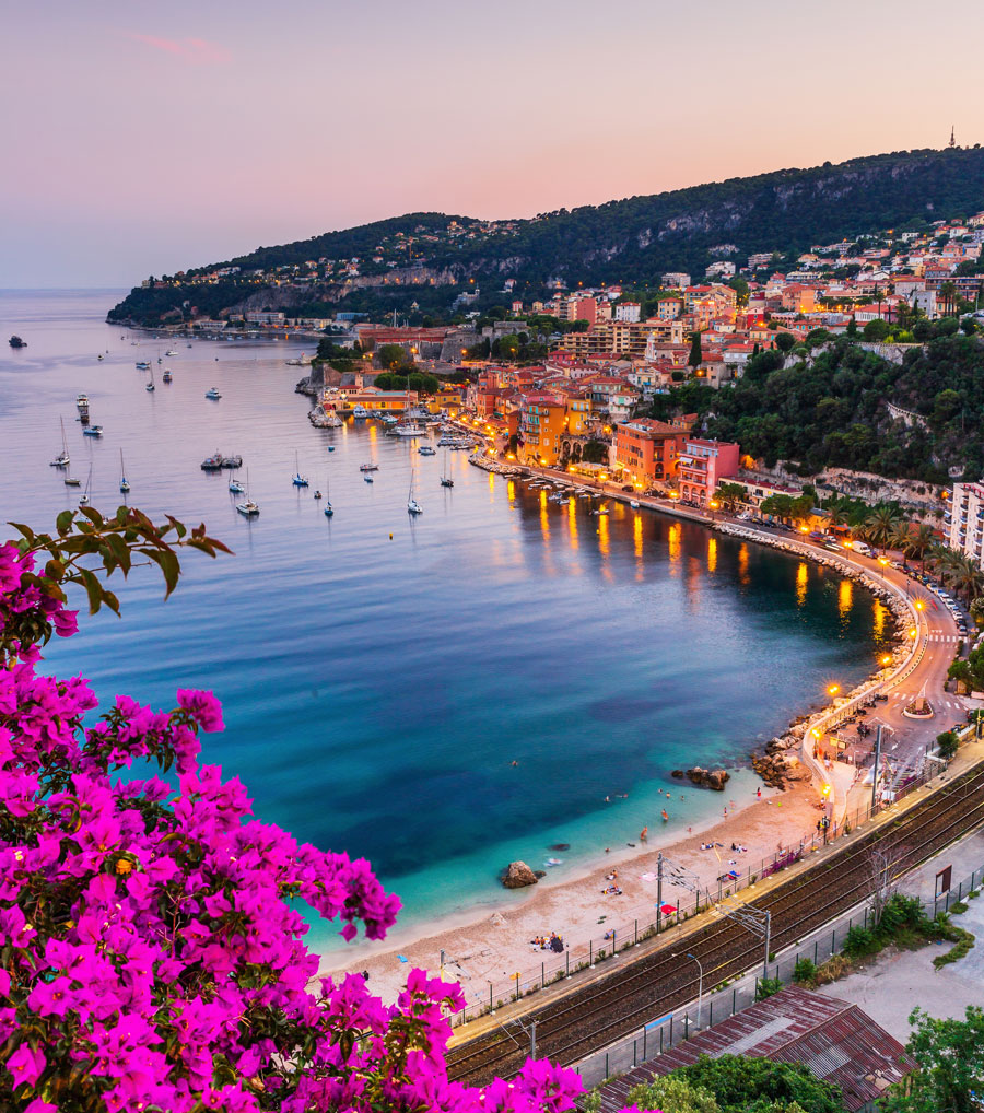 Uma cidade litorânea com uma baía curva ao pôr do sol. Prédios se alinham à beira da água, barcos estão ancorados na baía e flores rosa vibrantes estão em primeiro plano. Uma estrada e uma ferrovia correm ao longo da costa, levando a um cume panorâmico perfeito para os parceiros explorarem juntos.