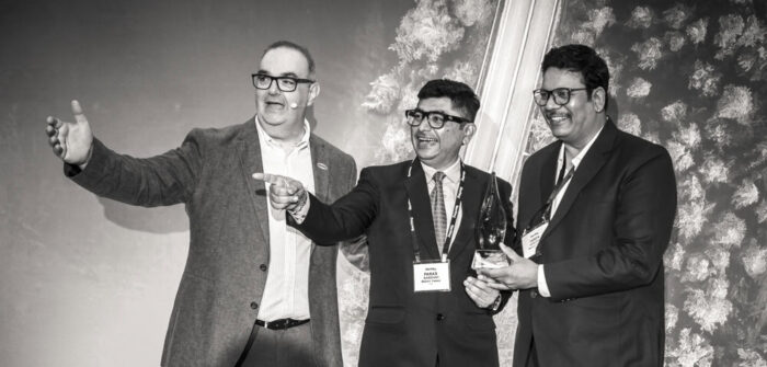 Three men in suits and glasses stand together, smiling and gesturing, with one holding a trophy. Adorned with conference badges, they are positioned against a backdrop featuring a forest and road, celebrating their achievement in the Partner Spotlight for Mahavir Traders.