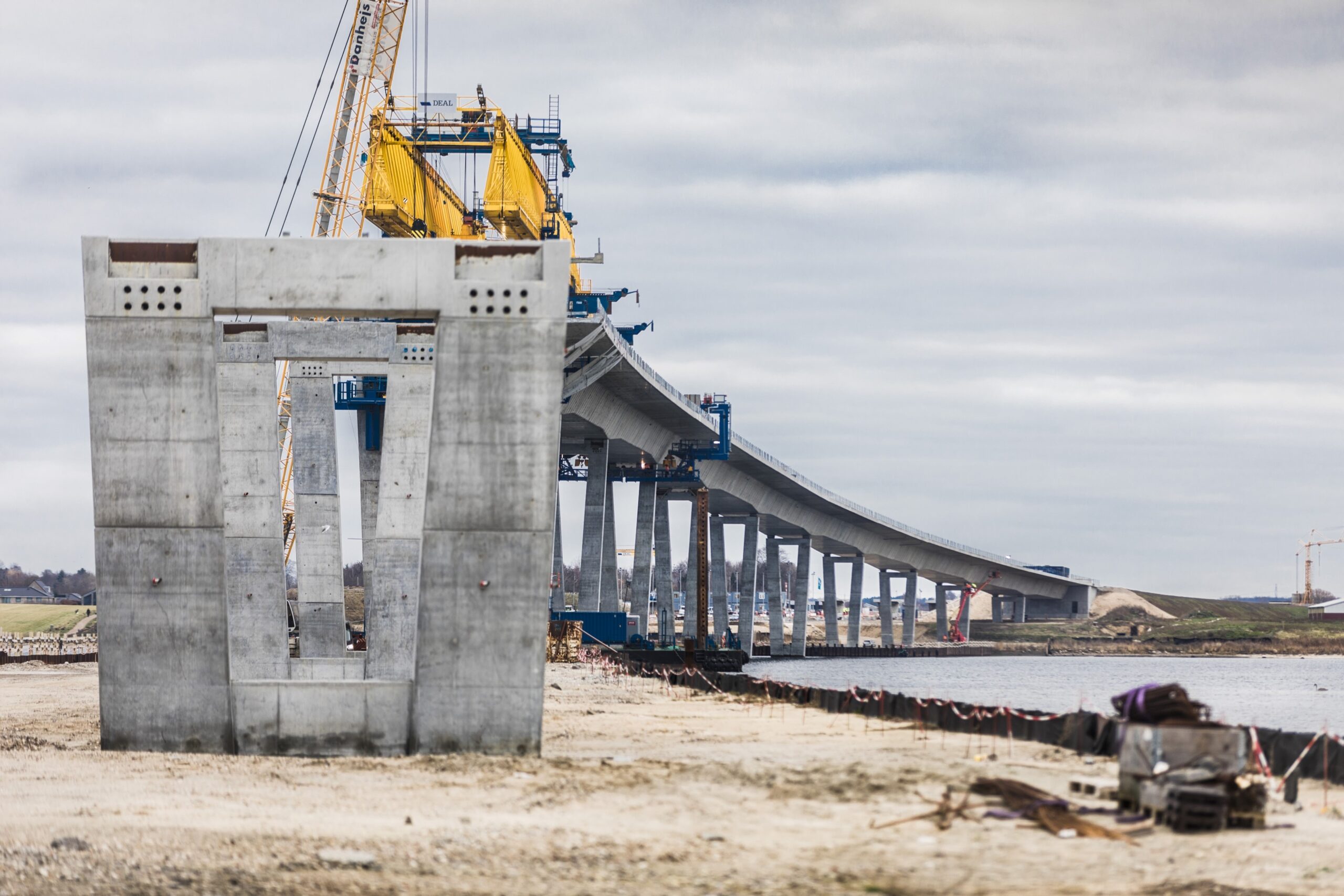 A large concrete section of a bridge in construction, orchestrated with meticulous construction planning, features cranes and other equipment. The bridge, overseen by the Danish Road Directorate, gracefully extends over water and land in the background.