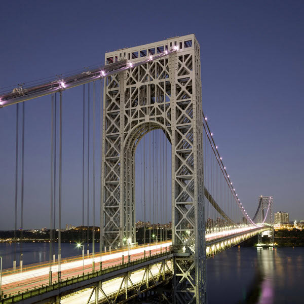 Eine beleuchtete Hängebrücke, die von der Hafenbehörde von New York unterhalten wird, überspannt in der Abenddämmerung ein Gewässer, im Hintergrund sind die Lichter der Stadt zu sehen.