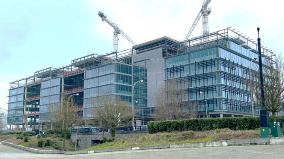 A modern, unfinished glass and steel building with two cranes on top stands tall. The background features a cloudy sky, while the foreground showcases a paved area with surrounding greenery and streetlights. The site has an air of meticulous planning, almost as if rendered from an Auto Draft blueprint.