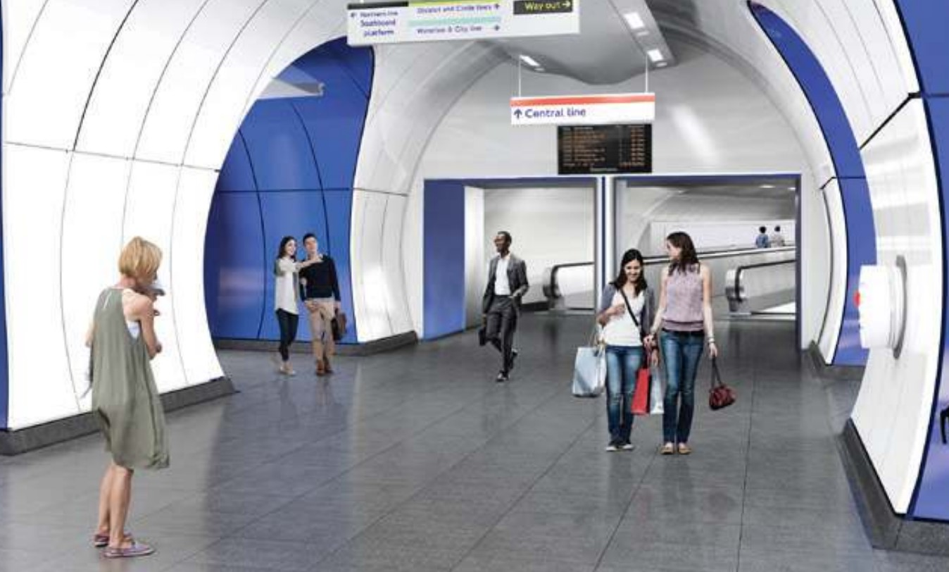 People walk through a modern underground station with blue and white walls and overhead signage indicating directions in the recently completed Dragados Subterranean Station Upgrade for the London Underground. Some individuals are holding bags while moving through the corridor.