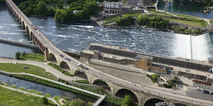 Aerial view of a historic stone arch bridge with strong, weathered columns stretching over a river, surrounded by lush greenery and nestled among industrial buildings nearby.