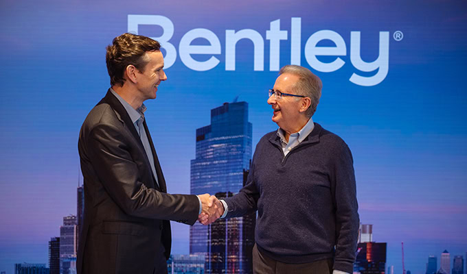 Zwei Männer schütteln sich die Hände vor einem großen Bildschirm mit dem Bentley-Logo und einer urbanen Skyline im Hintergrund.