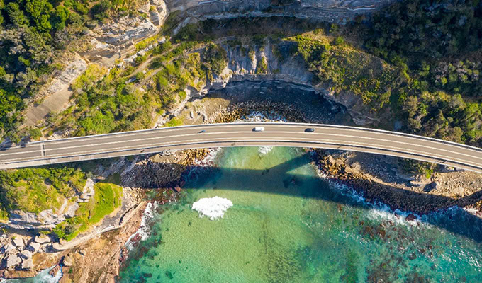 Aerial view of a coastal highway bridge showcasing sustainable engineering over clear blue-green water, with surrounding rocky shoreline and lush greenery.