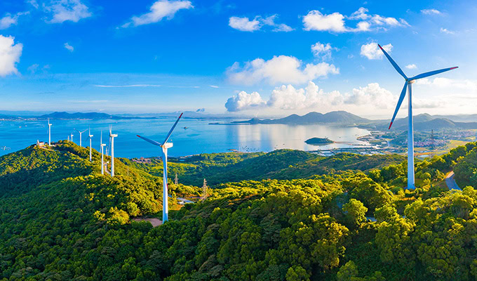 Wind turbines on a green hillside overlooking a bay, with scattered clouds and blue sky, offer insights into sustainable energy's beauty.