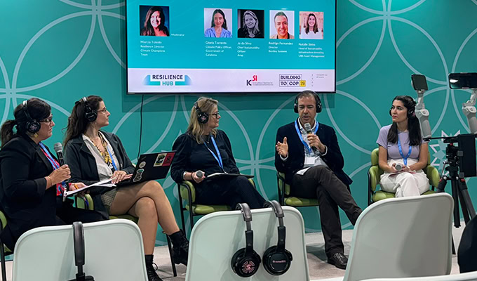 Five people seated and wearing headphones participate in a panel discussion. A screen behind them displays their names and photos.