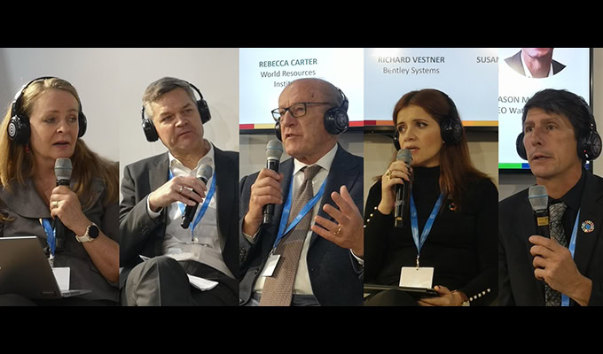 Five people sitting and holding microphones during a panel discussion, wearing headsets. Text with names and affiliations visible on the screen behind them.