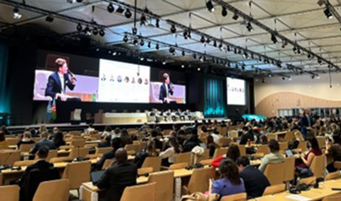 Large conference room with attendees seated at rows of desks, facing a stage with large screens displaying a speaker and panel information.