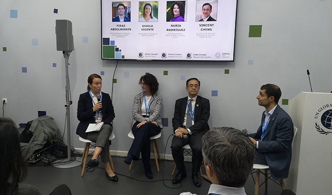 Four panelists are seated in a row on stage at a conference. A woman on the left is speaking into a microphone, while the others listen. A presentation slide with names and faces is displayed behind them.